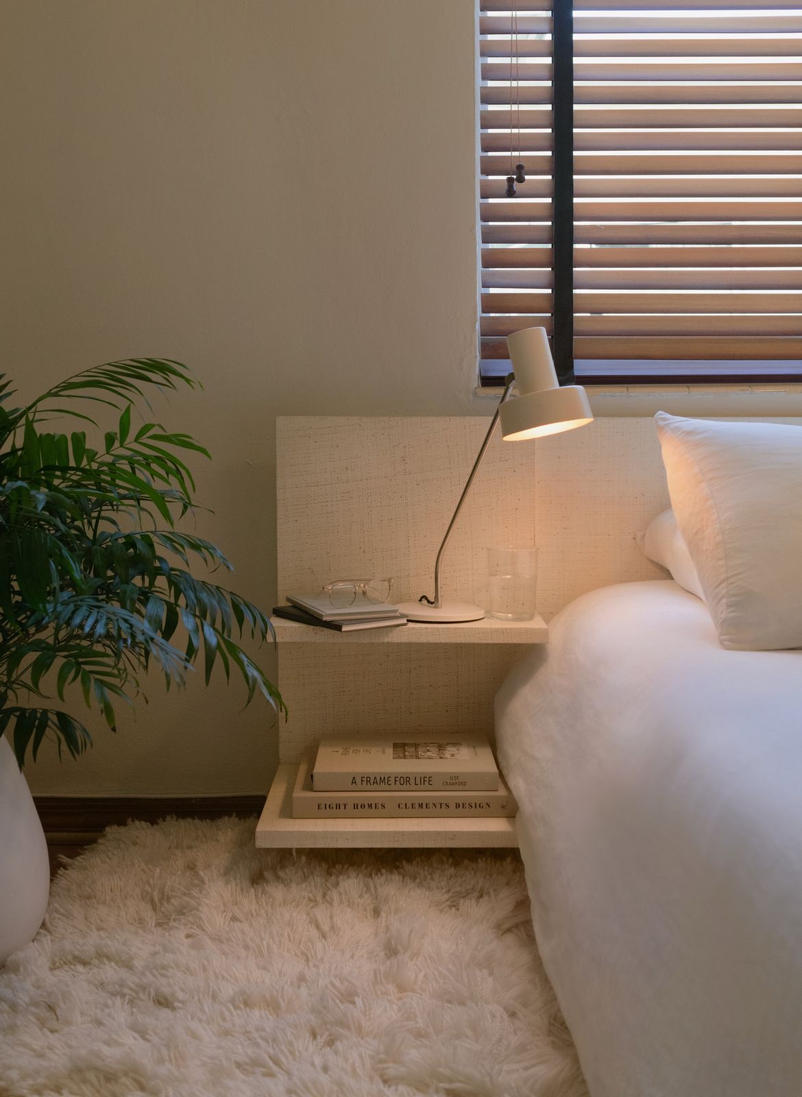 A sunlit bedroom by Studio DEA: white linen, a plaster side table, a palm and a shaggy rug