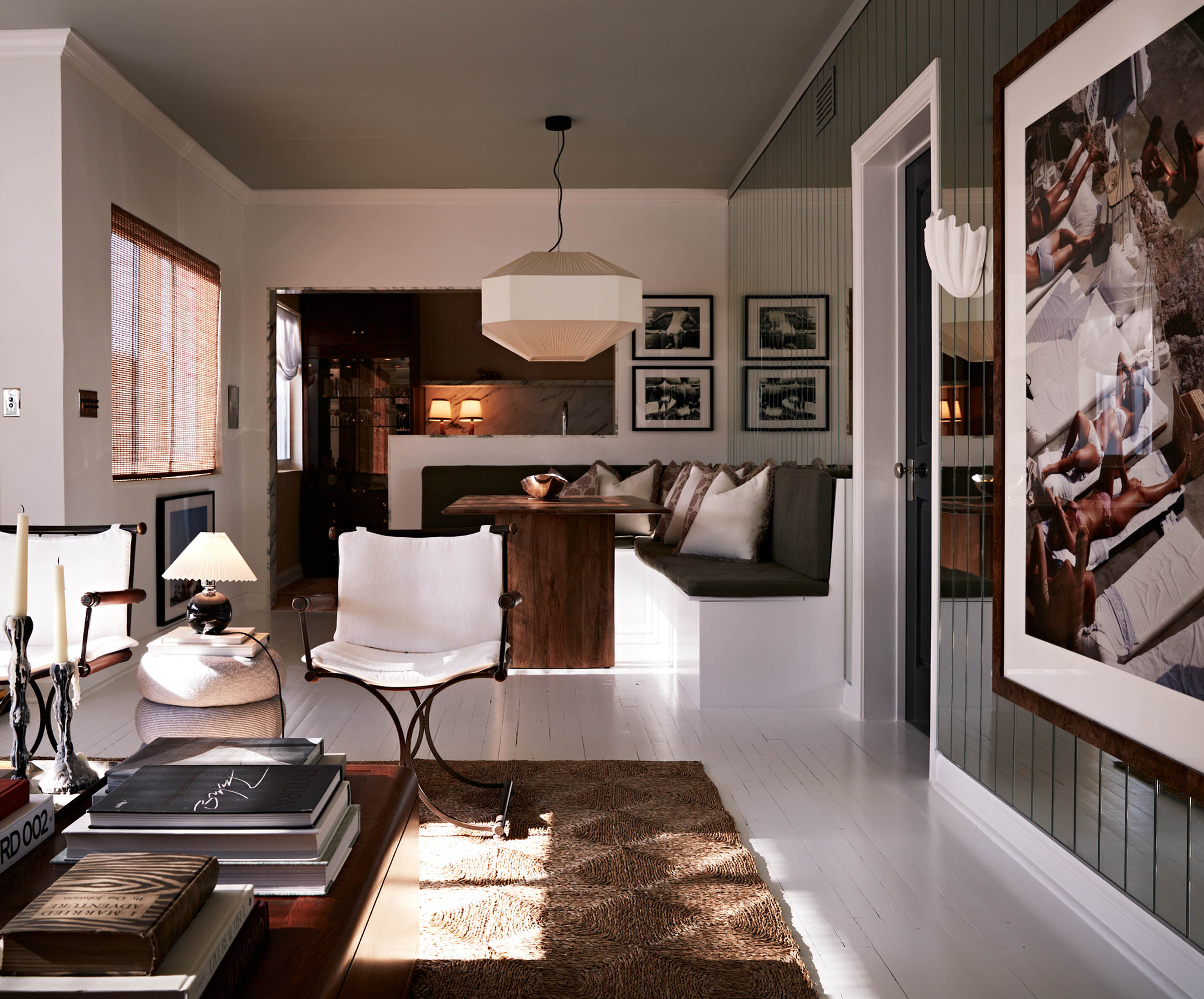 A living room by Studio DEA in late sun: a pleated pendant over a banquette, black-and-white photographs, a sling chair on a woven rug